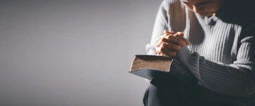 A person in a thoughtful pose, holding an open book while praying.