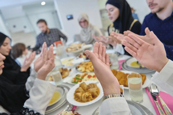 A group of people with hands raised in prayer around a dining table filled with food.