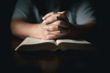 Hands clasped in prayer over an open book, possibly a Bible, on a wooden surface.